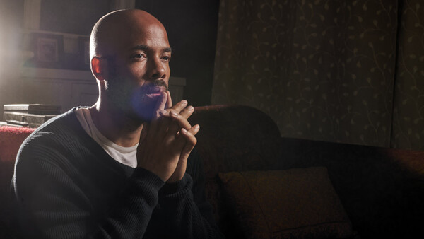 Man in dark long-sleeve shirt sitting indoors with hands clasped near face, looking pensively to the side.