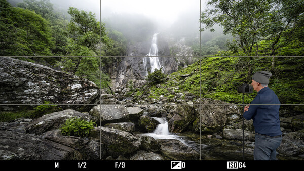 Photographer standing on mossy rocks beside a flowing stream with a misty waterfall visible in the forested background.