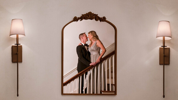 Couple in formal attire kissing on a staircase, reflected in an ornate arched mirror flanked by wall sconces.