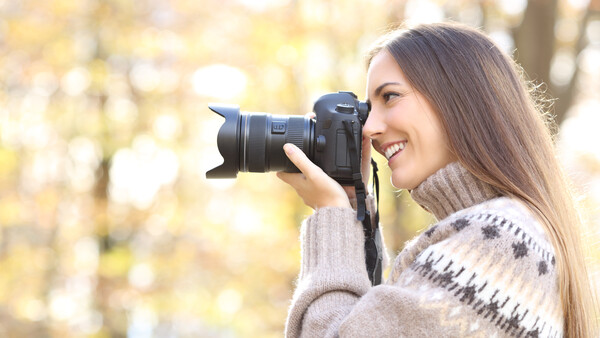 Woman with long brown hair holding a DSLR camera up to her face, smiling while framing a shot outdoors.