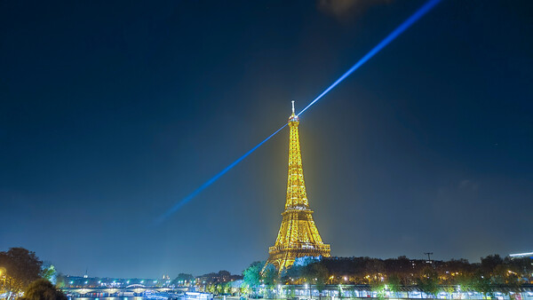 Eiffel Tower illuminated at night with a blue laser beam shooting upward into the dark sky, reflected in the Seine River below.