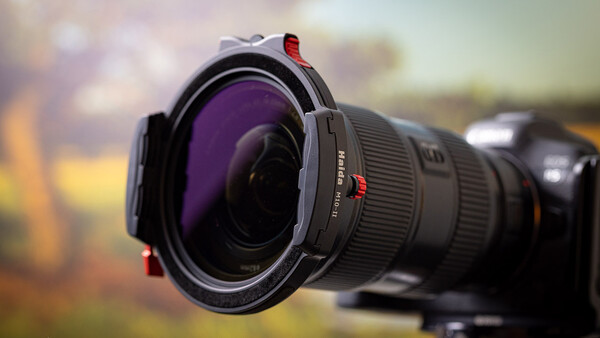 Wide-angle lens with purple filter mounted on camera body against blurred outdoor background.