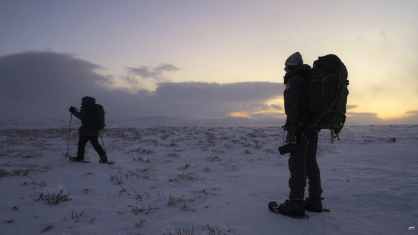 Three hikers with large backpacks trek across a snowy landscape during twilight.