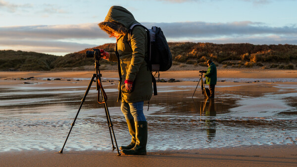 Photographer in golden jacket and backpack shooting at tripod on wet beach at golden hour.