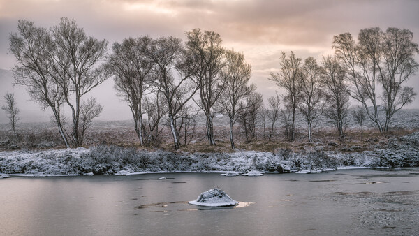 Bare trees line a snowy riverbank with a small ice-covered island in the foreground and misty hills beyond.