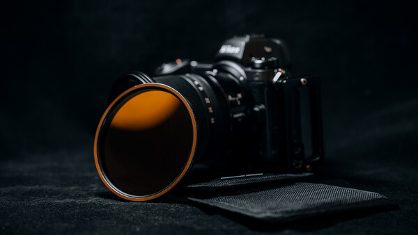 Camera lens with orange rim positioned in front of a DSLR body against a dark textured surface.