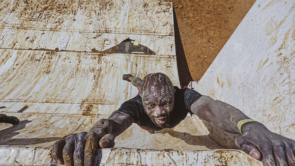 Mud-covered person climbing over a white wooden obstacle wall during an outdoor endurance event.