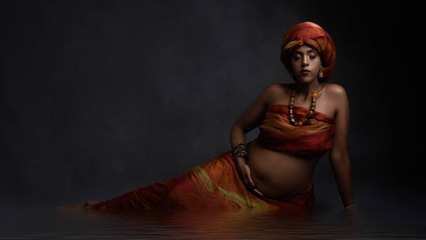 Woman in red-gold costume reclining in shallow water against dark background.