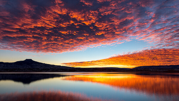 Dramatic sunset sky reflected in still lake water with mountain silhouette.