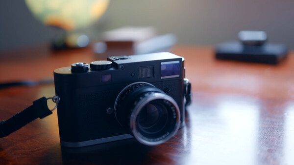 Black rangefinder camera with leather strap on wooden desk with blurred photography equipment in background.