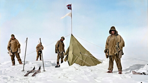 Five people in tan cold-weather gear standing around a canvas tent with a flag on a snowy landscape.