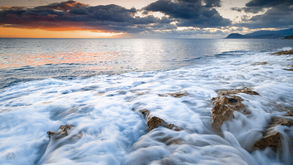Waves crash over rocky beach at sunset with dramatic orange and blue clouds reflected in the water.
