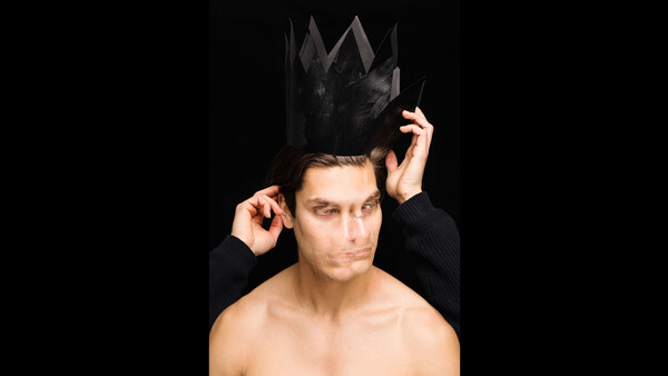 Shirtless man adjusting a black crown-shaped headpiece against a black background.