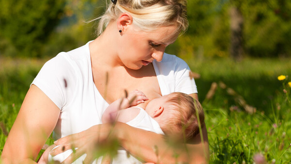 Mother breastfeeding infant outdoors in natural daylight surrounded by grass and greenery.