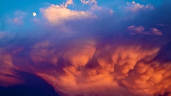 Dramatic cloudscape at dusk with towering cumulonimbus clouds illuminated in orange and gold against a deepening blue sky, with the moon visible above.