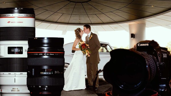 Wedding couple embracing under a modern dome structure, photographed through an array of Canon lenses and camera equipment in the foreground.