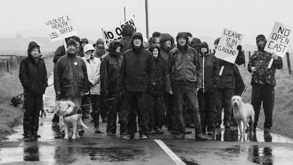 Group of protesters holding signs and walking along a wet road in overcast conditions.