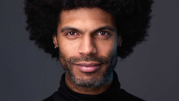 Portrait of a man with natural afro hair and facial hair against a neutral gray background.