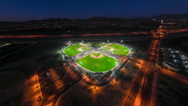 Aerial nighttime view of illuminated sports complex with three rectangular fields surrounded by parking areas and city lights.
