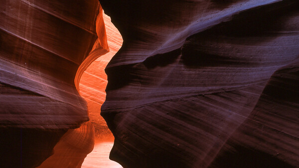 Slot canyon with undulating sandstone walls lit by warm orange light filtering from above, creating dramatic shadows and deep purple tones.