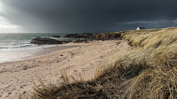 Dramatic coastal landscape with sandy beach, dune grasses, rocky outcrops, and stormy sky.