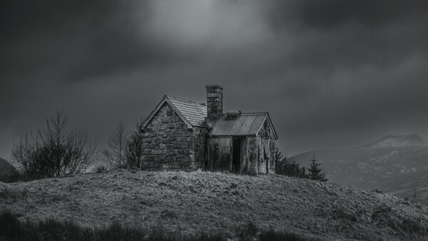 Abandoned stone house with deteriorating wooden additions sits isolated on a barren hilltop under dark storm clouds.