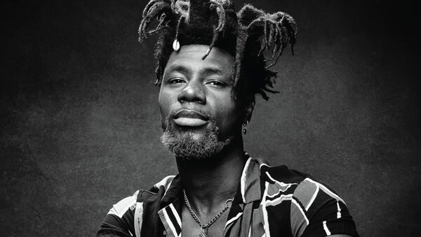 Black and white portrait of a man with styled dreadlocks, beard, and patterned shirt against dark background.
