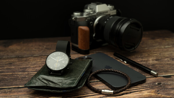 Minimalist flat lay of photographer's accessories on wood surface including analog camera, wristwatch, leather wallet, and cable.