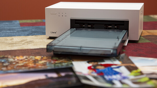 Compact photo printer with paper tray open, positioned over scattered printed photographs and colorful background.