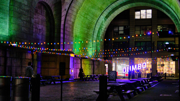 Historic stone archway illuminated with vibrant green, purple, and blue lights, decorated with warm-toned string lights.