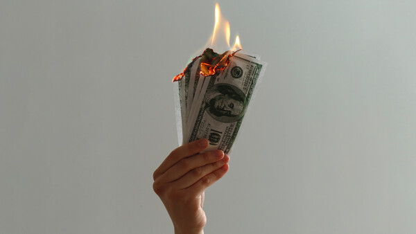 Hand holding burning U.S. dollar bills against a neutral background.