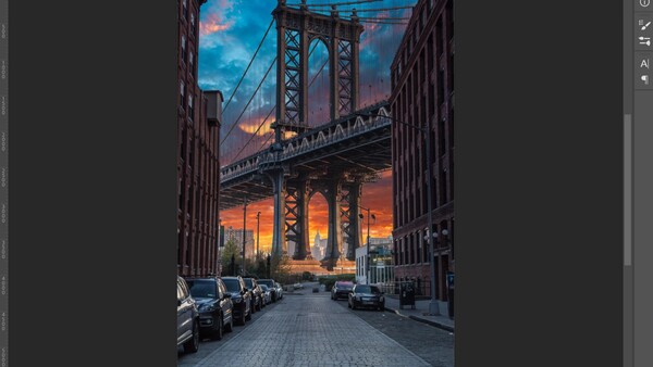 Urban street framed by buildings with suspension bridge and dramatic sunset sky at dusk.
