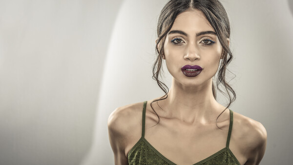Woman with dark wavy hair and burgundy lipstick wearing olive green camisole against neutral backdrop.