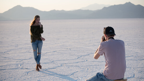 Male photographer seated on a stool captures a female model posing on a salt flat with mountains and water in the background.
