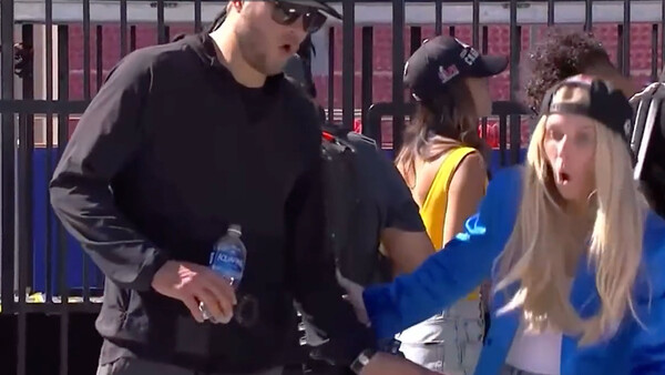 Group of people gathered outdoors behind a fence, with one man in black clothing and sunglasses conversing with others.