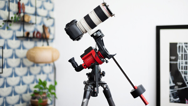 A telescope mounted on a red equatorial mount with a telephoto lens attached, positioned in an indoor studio setting.