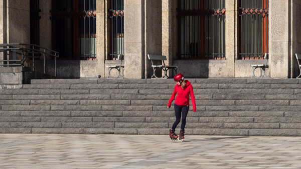 Person in red jacket rollerskating across a wide stone plaza in front of a modernist building.