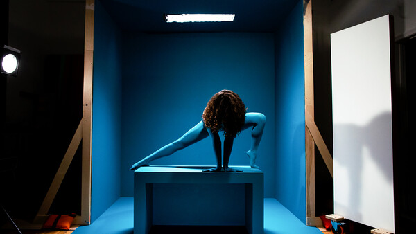 Person in deep forward fold pose on platform inside illuminated blue studio box with fluorescent overhead lighting.