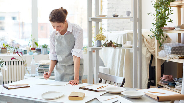 Person leaning over a desk arranging ceramic plates and printed materials in a bright studio space.