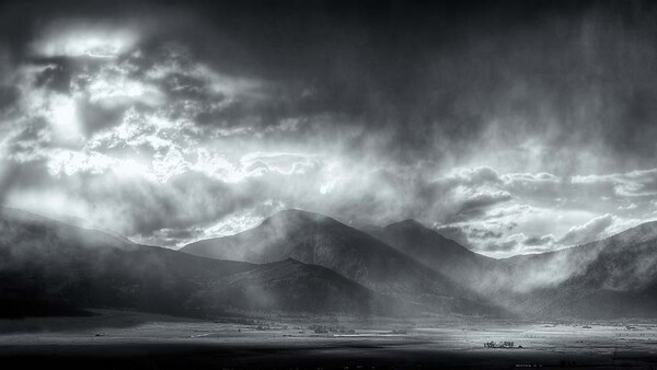 Dramatic black and white landscape with a solitary mountain rising from a vast plain beneath stormy clouds.