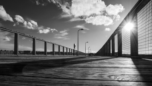 Empty concrete pathway between tall mesh fencing under bright sunlight and dramatic clouds.