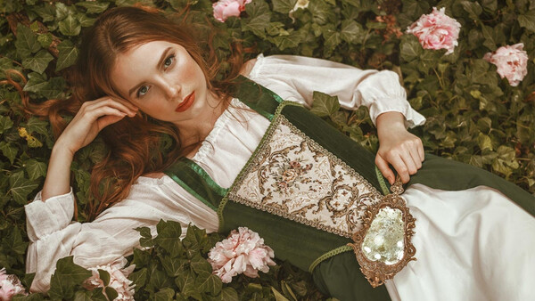 Woman in green dirndl dress reclining among pink roses and foliage.