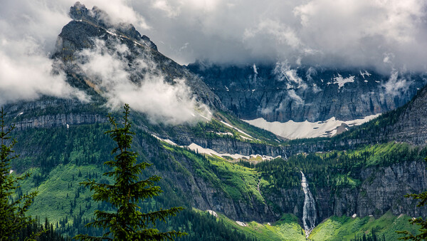 Alpine valley with forested slopes, a cascading waterfall, and dramatic peaks shrouded in low clouds.