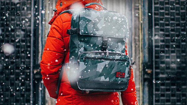 Person wearing red jacket and backpack standing in urban setting with falling snow.