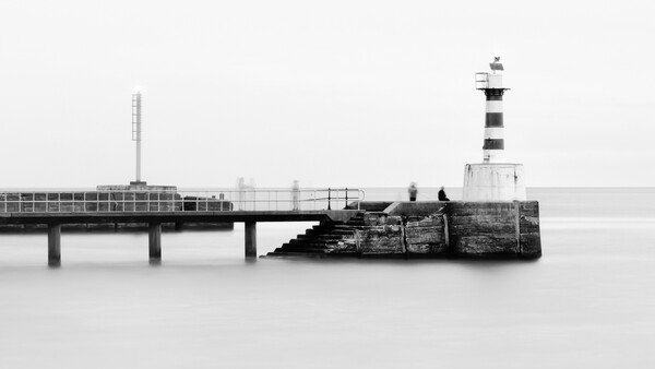 Lighthouse at the end of a long pier extending into calm water, captured in black and white.