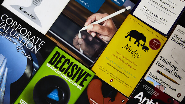 Hand holding a stylus over an open business book surrounded by stacked professional development books on a dark surface.