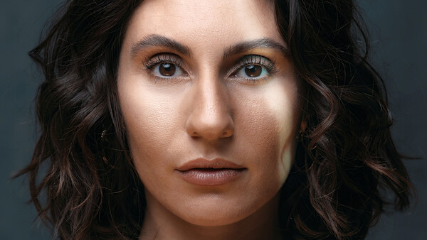 Close-up portrait of a woman with dark wavy hair and defined eye makeup against a neutral gray background.