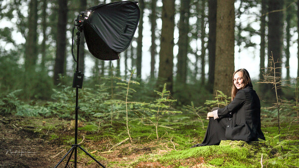 Woman in black blazer seated in forest clearing with professional studio light on stand.
