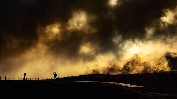 Silhouetted figures standing by a fence with dramatic dust and smoke billowing across a dark sky.
