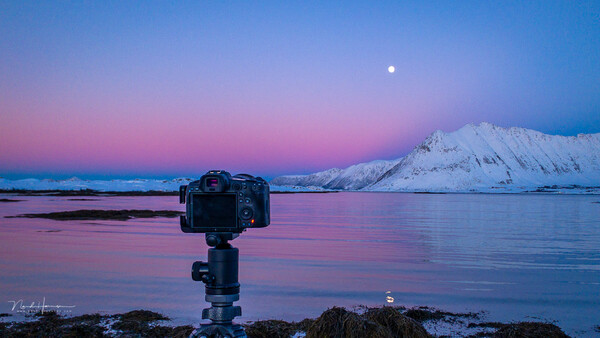 Camera on tripod mounted on rocky shore facing snow-capped mountain across still water at twilight with moon visible.